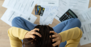 (Overhead view of a stressed person clutching their head, surrounded by scattered bills, credit cards, and a calculator—symbolizing financial overwhelm and debt anxiety.)