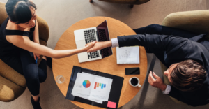 (Two professionals shaking hands across a round table with charts, a laptop, and coffee, signifying a successful financial or business agreement.)